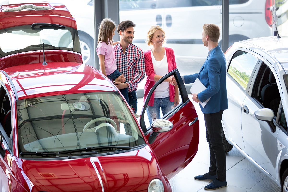 Smiling car agent showing vehicle to young family, all stand together next to the car in the car dealership saloon